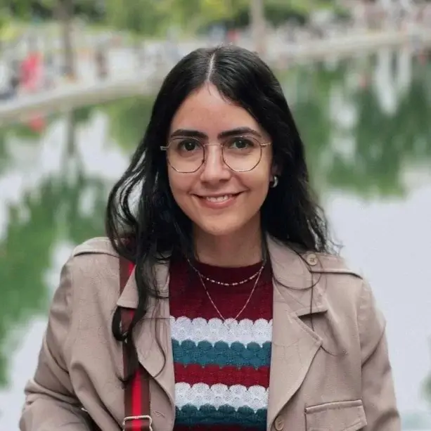 Maria está de óculos e blusa listrada na frente de um lago