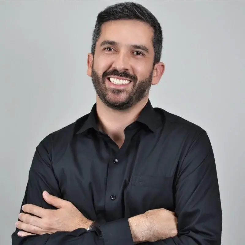 Homem branco de barba e camisa social preta com braços cruzados sorri