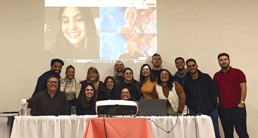 Capacitação Customer Experience com Weber e participantes do treinamento atrás de uma mesa e com projeção de reunião na parede