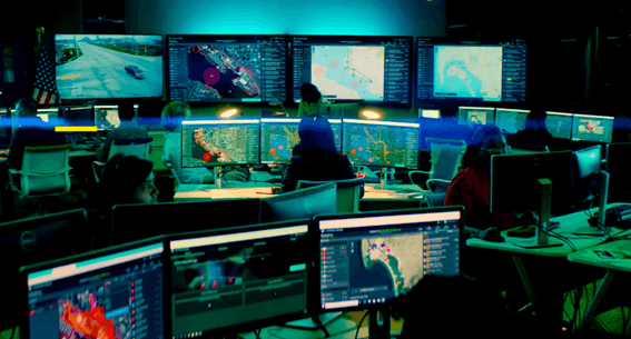 Centro de operações com múltiplos monitores exibindo mapas e dados geoespaciais em tempo real, utilizado para monitoramento e gestão de crises.