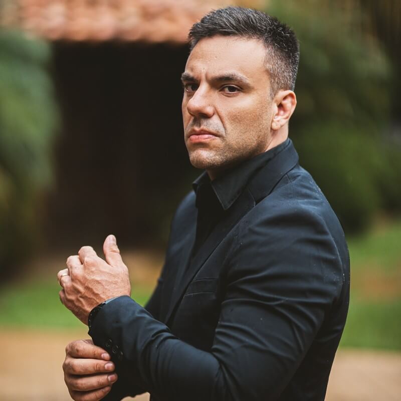 homem de terno escuro, olhando diretamente para a câmera, com expressão séria. Ele está ajustando a manga do paletó, em um ambiente externo com árvores ao fundo