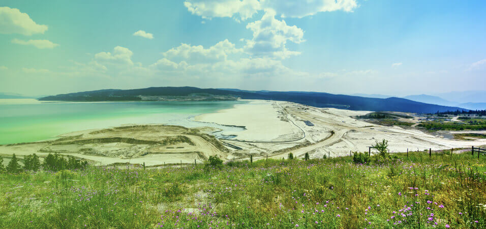 Vista panorâmica de uma área de mineração ao lado de um grande lago verde, com vegetação e flores silvestres no primeiro plano. A imagem destaca o impacto das operações de mineração na paisagem e a integração com o ambiente natural.