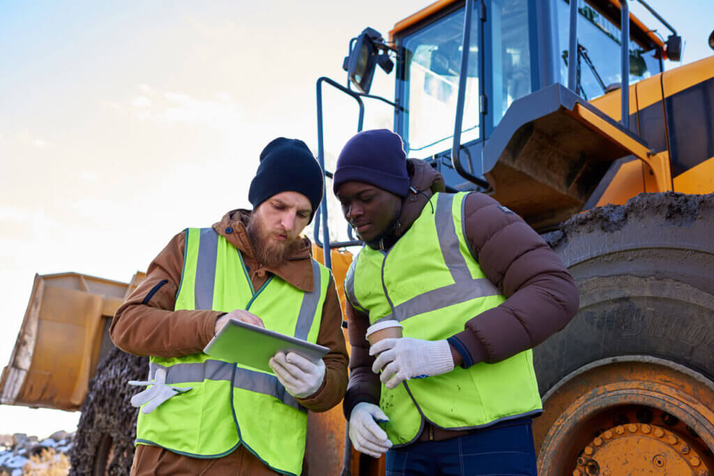 Dois trabalhadores de mineração, vestindo coletes de alta visibilidade e tocas, analisam dados em um tablet em frente a um grande maquinário de mineração. Ambos estão focados, destacando o uso de tecnologia para melhorar operações no setor mineral.
