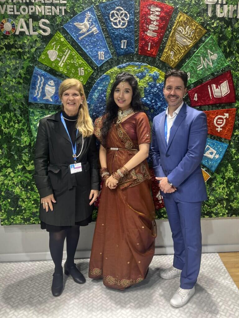 Marlos Batista posando ao lado de duas participantes da COP 29, em frente a um painel decorativo que destaca os Objetivos de Desenvolvimento Sustentável (ODS) da ONU.