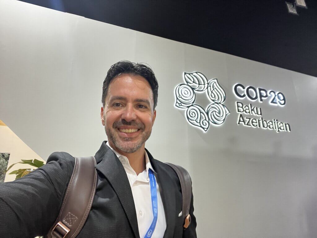 Marlos Batista, Diretor de Sustentabilidade da Imagem Geosistemas, sorrindo para a câmera em frente à logo oficial da COP 29 em Baku, Azerbaijão.