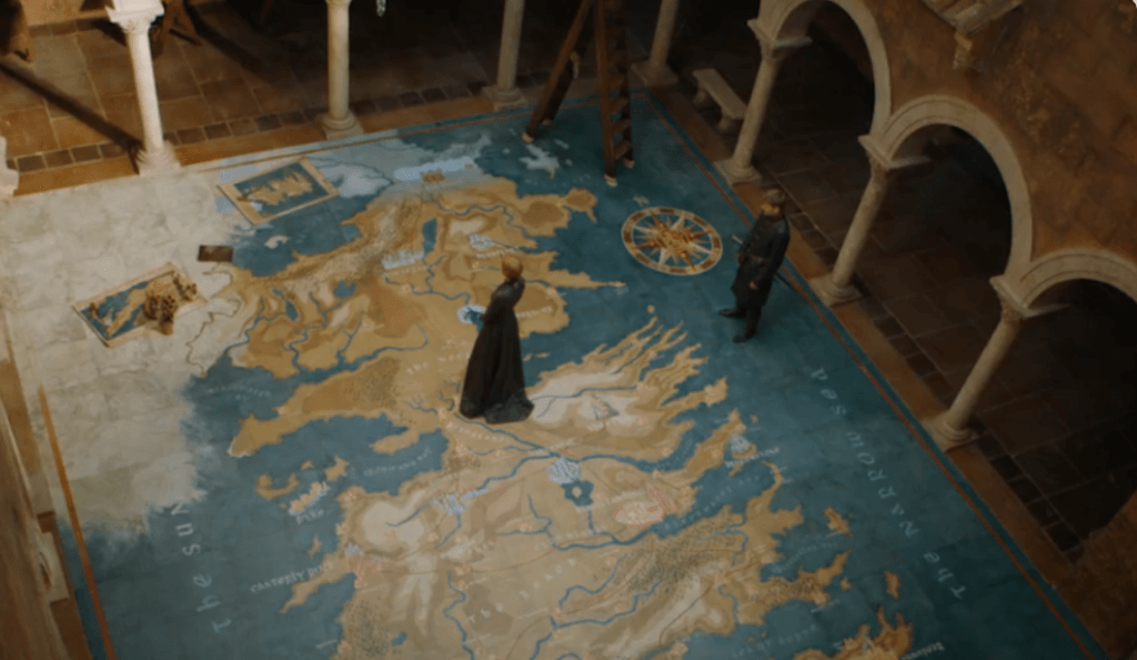 Cena de uma série com dois personagens em pé sobre um enorme mapa pintado no chão, representando um continente fictício com rios, montanhas e nomes geográficos. O local é uma sala ampla, com colunas, arcos e chão de pedra, iluminada suavemente.