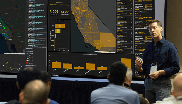 Palestrante apresentando um painel interativo de geoinformação, destacando dados visuais de mapas e gráficos de uma região, enquanto uma audiência observa atenta. A apresentação parece focar na análise de dados geoespaciais e indicadores regionais.
