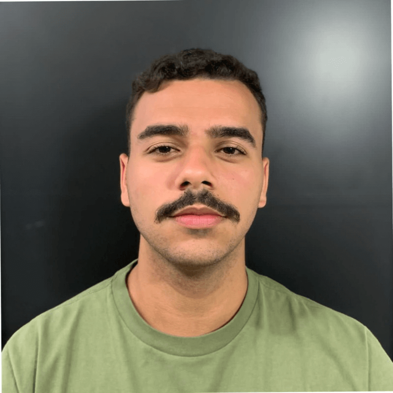 Retrato frontal de um homem jovem com cabelo curto e bigode, vestindo uma camiseta verde-oliva, posando contra um fundo preto liso.