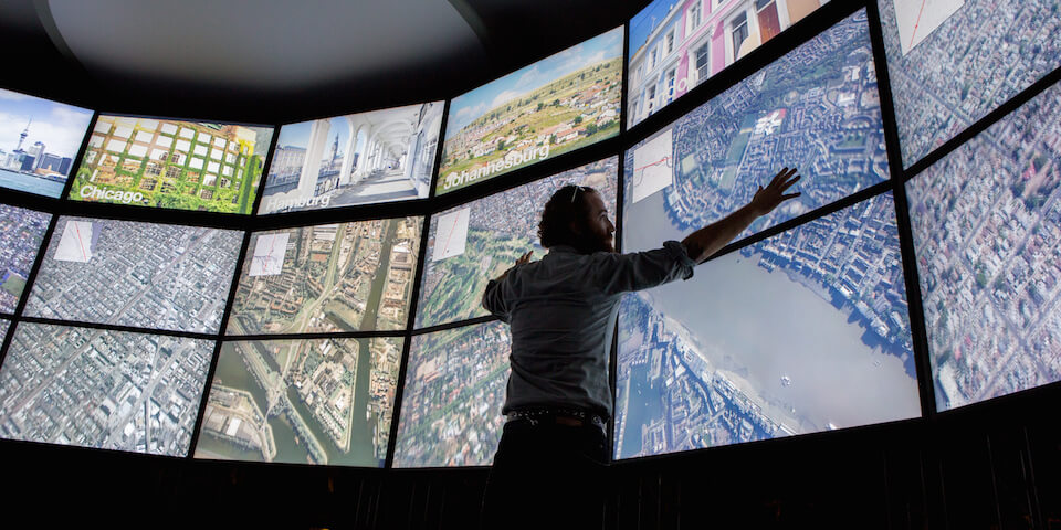 Pessoa interagindo com um grande painel de múltiplas telas exibindo mapas e imagens de satélite de diversas cidades ao redor do mundo, como Chicago, Hamburgo e Joanesburgo. A cena sugere monitoramento geoespacial avançado, possivelmente para análise de dados, planejamento urbano ou resposta a emergências.