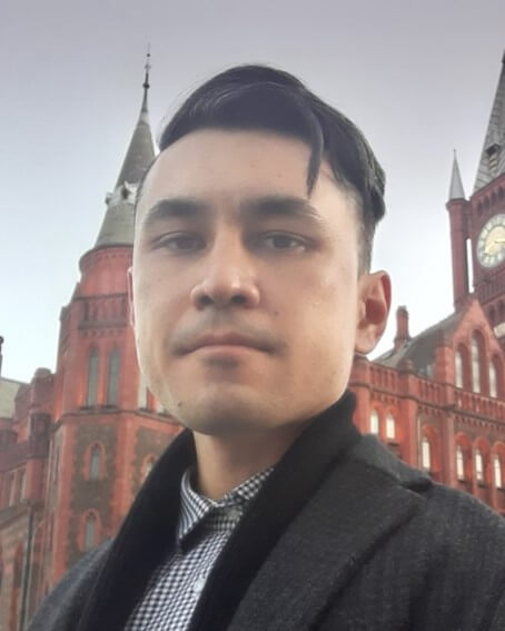 Um retrato de um homem jovem com cabelo curto e escuro, usando um casaco preto e uma camisa xadrez. Ele está posando para uma selfie ao ar livre, com um prédio de tijolos vermelhos e torres no fundo, possivelmente um edifício histórico ou universitário.