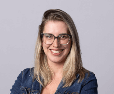 Mulher loira sorrindo, usando óculos de grau e jaqueta jeans em frente a um fundo neutro