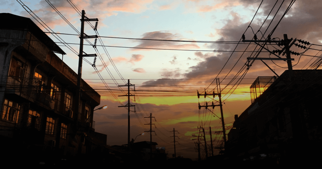 Silhueta de edifícios e postes de energia ao pôr do sol, destacando a infraestrutura elétrica em um cenário urbano.