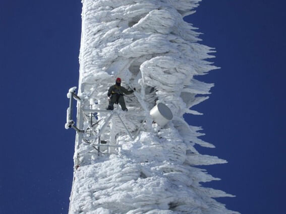 Trabalhador em uma torre de telecomunicações coberta de gelo, realizando manutenção em condições climáticas extremas.