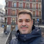 Homem de meia-idade, com cabelos castanhos curtos e barba rala, sorrindo em ambiente urbano. Ele usa casaco escuro e aparece em frente a um edifício de estilo europeu com varandas de ferro e janelas simétricas. Foto ao ar livre, provavelmente em cidade europeia.