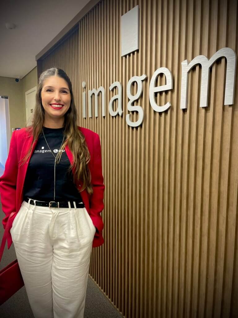 Aline Coimbra, Analista de Sucesso do Cliente da Imagem Geosistemas, posa sorridente em frente ao logotipo da empresa
