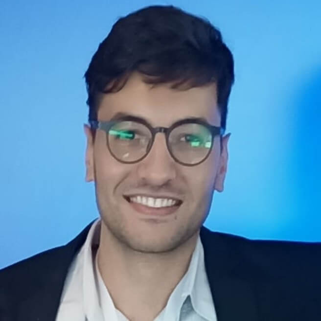 Pessoa sorridente usando óculos e blazer, com fundo azul suave.