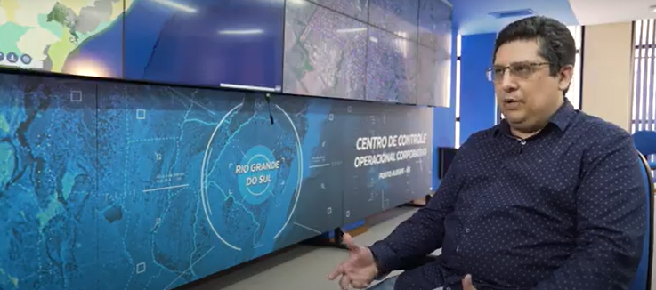 Profissional da CORSAN fala sobre o projeto Smart Water com ArcGIS, em frente ao Centro de Controle Operacional da companhia no Rio Grande do Sul, com painel de mapas ao fundo.