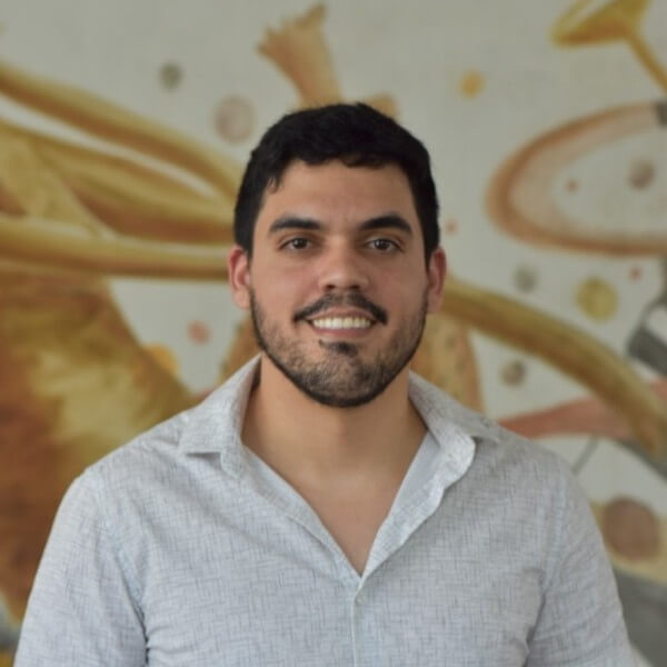 Retrato de um homem sorridente, com barba curta e camisa clara, posando em ambiente interno com mural artístico ao fundo.