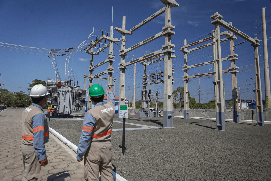 Técnico aponta para poste com transformador e fiação em área urbana, enquanto outro colaborador realiza manutenção com apoio de caminhonete.