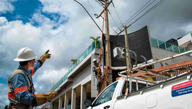 Técnico aponta para poste com transformador e fiação em área urbana, enquanto outro colaborador realiza manutenção com apoio de caminhonete.