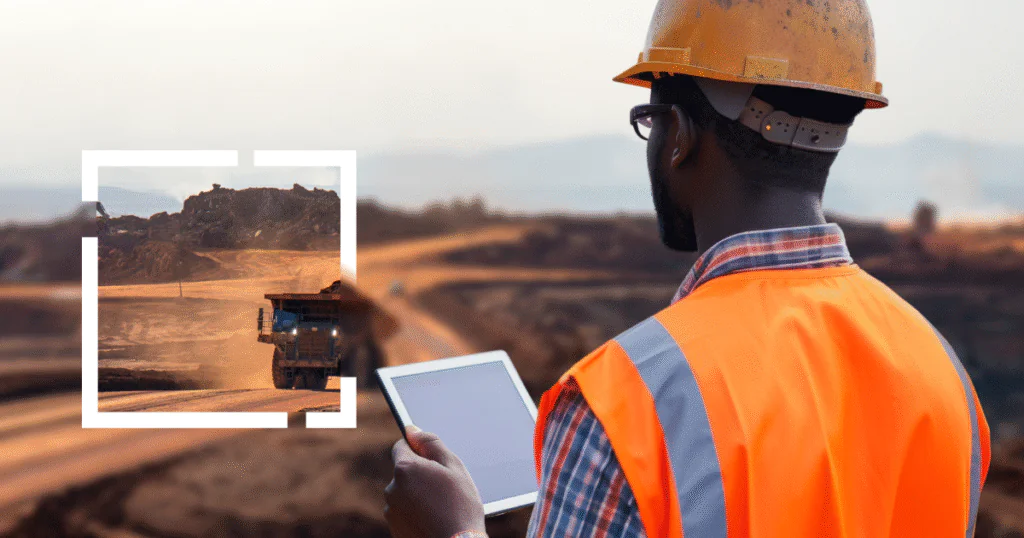 Trabalhador de mineração com colete laranja e capacete, usando tablet para monitorar operações em uma mina a céu aberto com caminhão basculante ao fundo