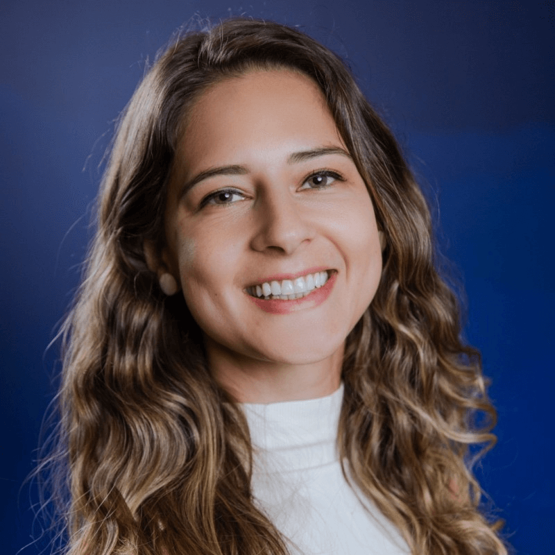 Retrato de uma mulher jovem sorridente com cabelos longos e ondulados castanhos, usando blusa clara de gola alta. O fundo é azul em degradê, destacando o rosto iluminado e o clima profissional da foto.