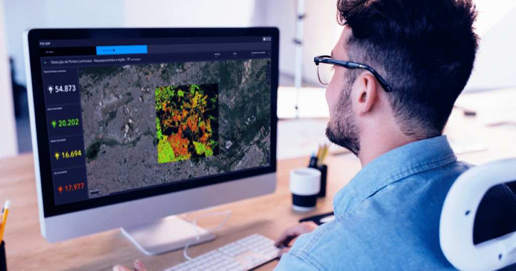 Homem jovem utilizando óculos, visto de trás, trabalha em um computador de mesa com tela grande exibindo um mapa temático colorido e um painel com estatísticas de pontos luminosos urbanos, sugerindo análise geoespacial de iluminação pública em ambiente moderno de escritório.